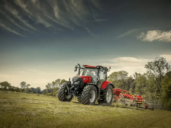 Rød traktor fra Case IH Maxxum-serien med høyvender i arbeid på en solfylt eng.