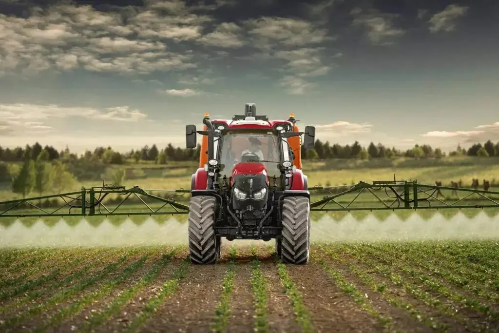 Rød traktor sprøyter en åker med lange bommer under en skyet himmel.