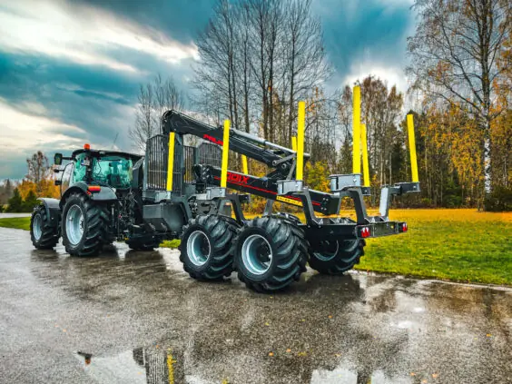 En mørk traktor med en PALMS HMWD 3.2 tømmervogn parkert på våt asfalt.