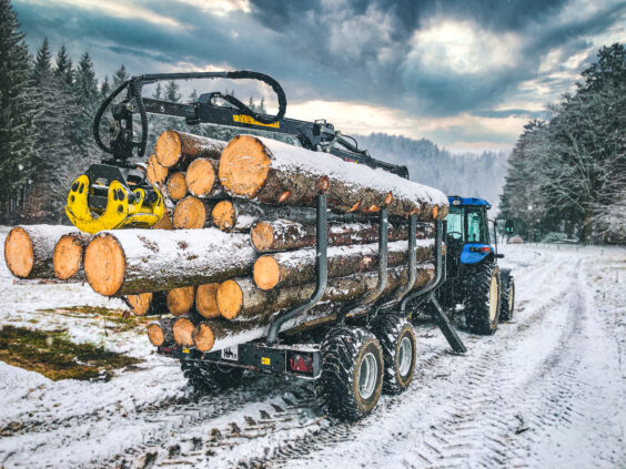 PALMS 8SX tømmervogn lastet med tømmerstokker trukket av en blå traktor i snøen.
