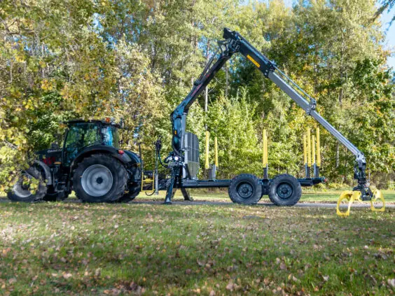 En traktor med PALMS 7.87 skogskran og tømmervogn stående foran grønne trær.