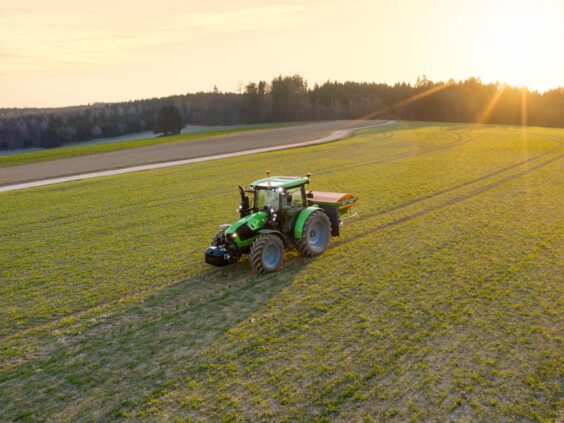 Grønn Deutz-Fahr Series 5 traktor sprer gjødsel på en grønn åker i solnedgang.