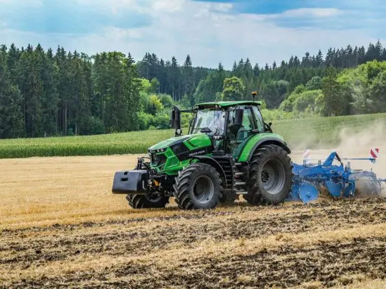 En grønn Deutz-Fahr Series 6 traktor som pløyer en åker med skog i bakgrunnen.