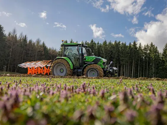 Grønn Deutz-Fahr Series 6C traktor pløyer en åker ved en skog.