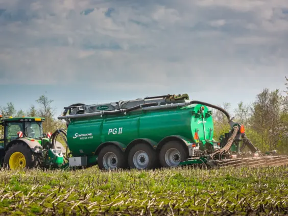 En grønn traktor som trekker en SAMSON PG II gjødselvogn på en åker.
