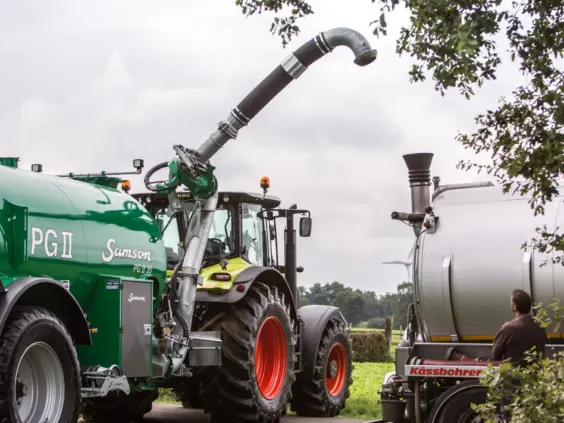 En grønn SAMSON PG II gyllevogn koblet til en traktor under fylling fra en tankbil.