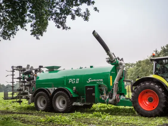 En grønn SAMSON PG II gjødselvogn trukket av en traktor på et jorde.