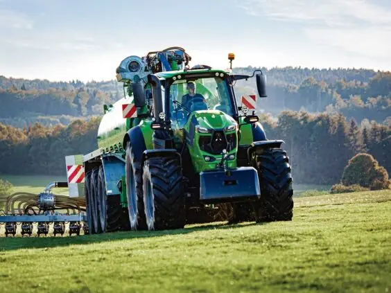 Grønn Deutz-Fahr Series 8 traktor med gjødselvogn i arbeid på en solfylt åker.