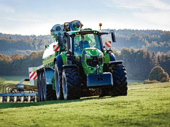 Grønn Deutz-Fahr Series 8 traktor med gjødselvogn i arbeid på en solfylt åker.