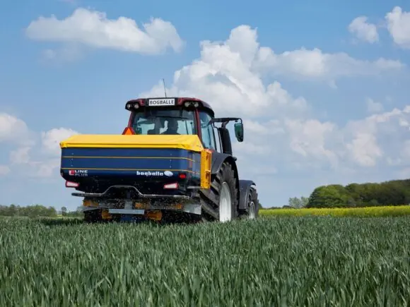 En traktor i arbeid med en Bogballe L20 plus kunstgjødselspreder på en grønn åker.