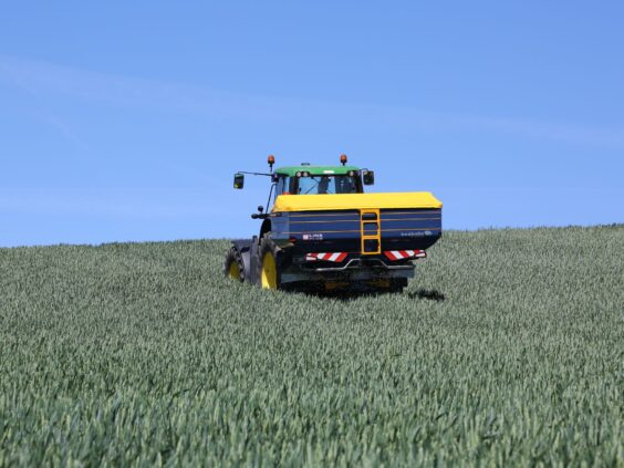En traktor med Bogballe M35 plus kunstgjødselspreder i en grønn kornåker under blå himmel.