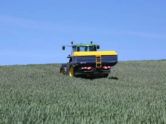 En traktor med Bogballe M35 plus kunstgjødselspreder i en grønn kornåker under blå himmel.