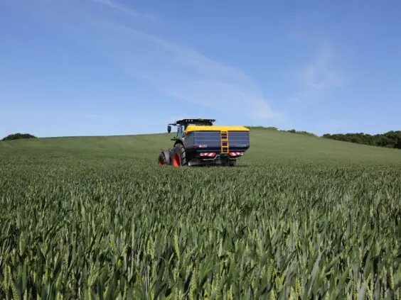 En traktor med Bogballe M45W plus kunstgjødselspreder i en grønn åker under blå himmel.