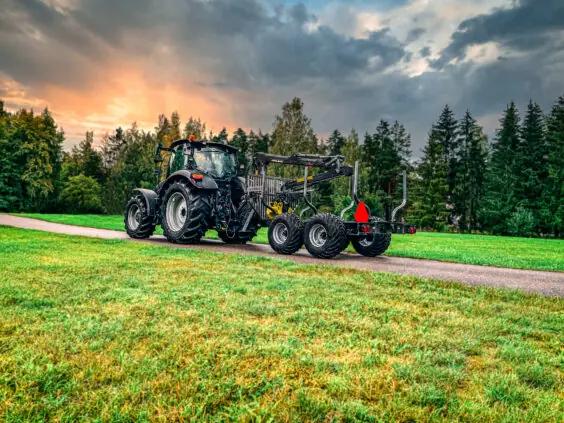 Svart traktor med PALMS 6S tømmervogn på en landevei under en fargerik solnedgang.