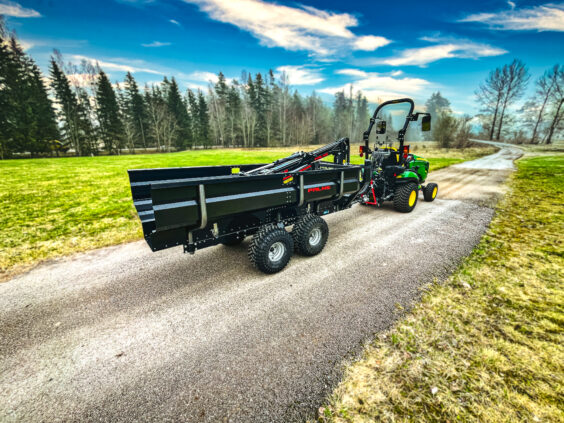 En PALMS 2D tømmervogn koblet til en grønn traktor på en grusvei i naturen.