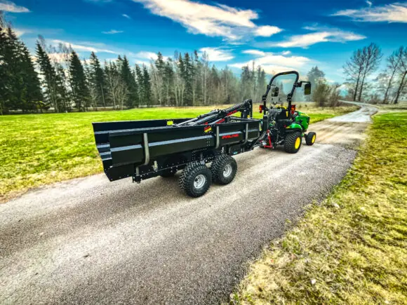 En PALMS 2D tømmervogn koblet til en grønn traktor på en grusvei i naturen.