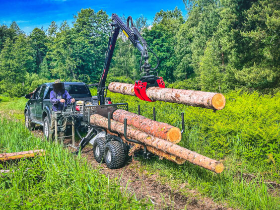 En PALMS 2D tømmervogn laster tømmerstokker bak en pickup i skogen.