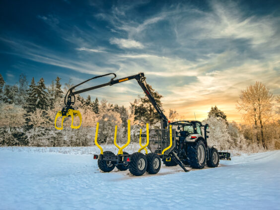 Traktor med PALMS 8SX tømmervogn på snødekket vei i et vakkert vinterlandskap under solnedgang.