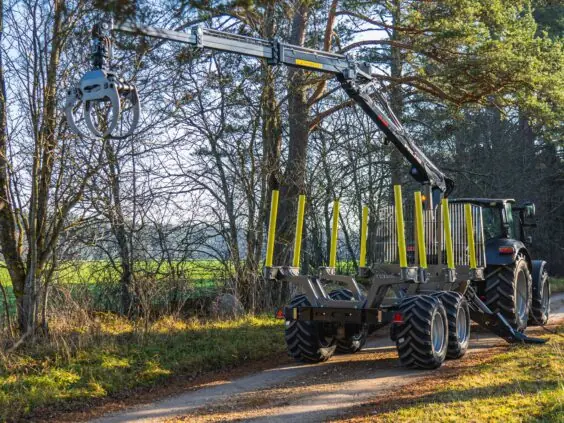 Traktor med PALMS 5.85 skogskran og tømmervogn på en skogsvei i solskinn.