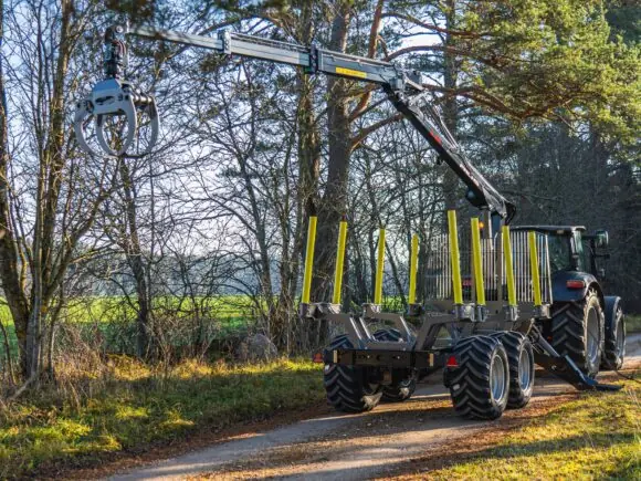 Traktor med PALMS 5.85 skogskran og tømmervogn på en skogsvei i solskinn.