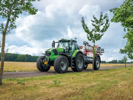 Grønn Deutz-Fahr Series 6 traktor med tilhengersprøyte kjører på en landevei mellom jorder.