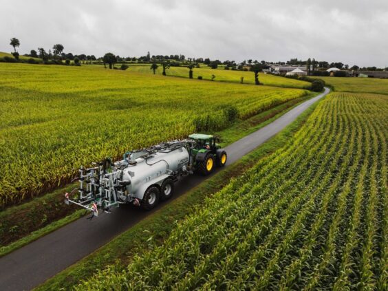Grønn traktor som trekker en Pichon SV-serien vakumvogn langs en vei gjennom store maisfelt.