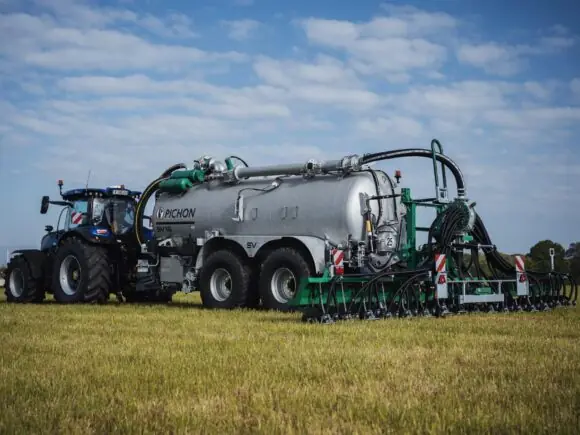 En blå traktor som trekker en Pichon SV-serien vakumvogn på en gresskledd mark.