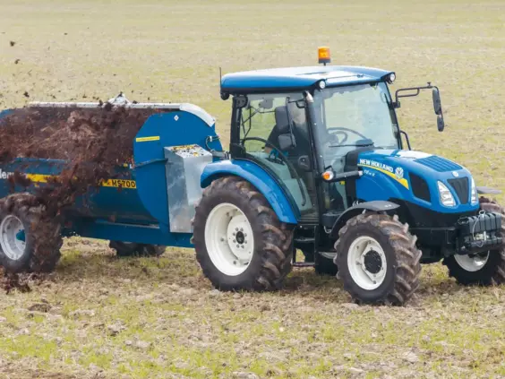 En New Holland T4S traktor trekker en blå gjødselspreder på en åker.