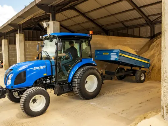 En blå New Holland Boomer 25 C-55 mini traktor med tilhenger i en lagringshall.