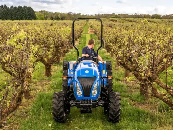 Blå mini traktor Boomer 25 C-55 som kjører mellom frukttrær i en grønn frukthage.
