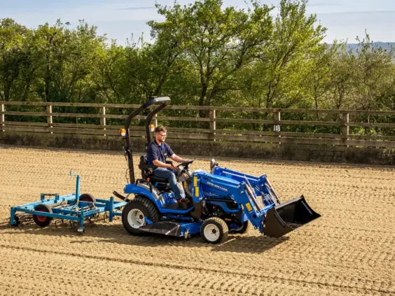 En blå New Holland Boomer 25 C-55 mini traktor i arbeid på en sandbane.