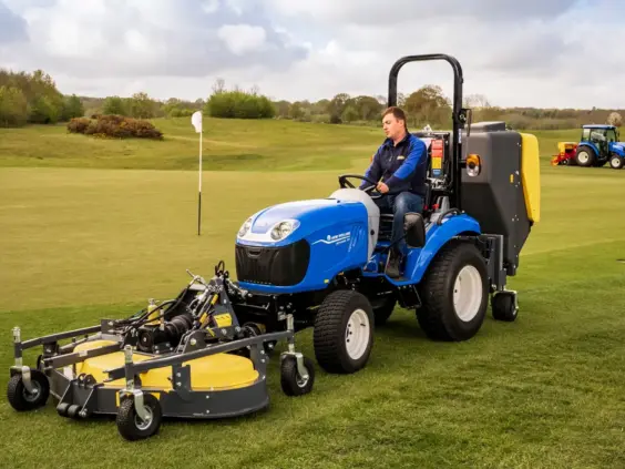 Blå New Holland Boomer 25 C-55 mini traktor med gressklipper på en grønn golfbane.