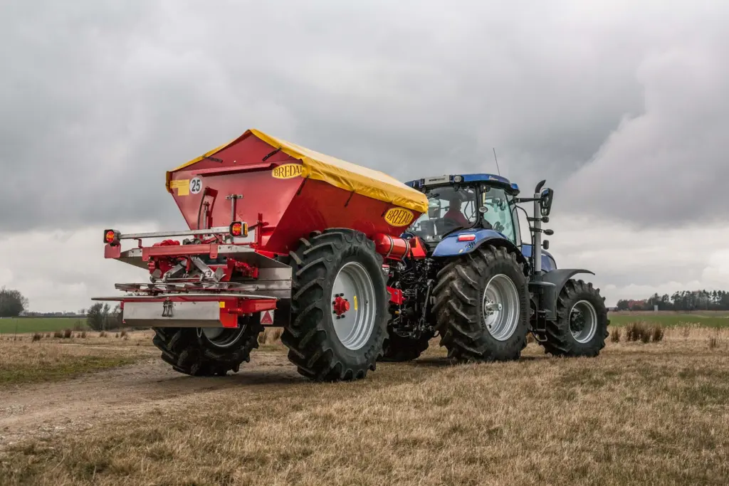En blå traktor på et jorde med en rød Bredal kalkspreder montert bak.