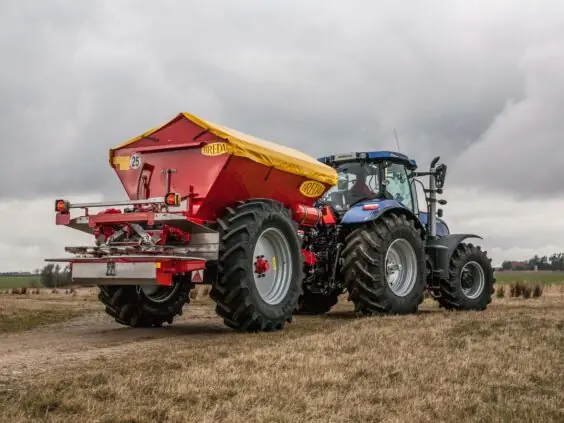 En blå traktor på et jorde med en rød Bredal kalkspreder montert bak.