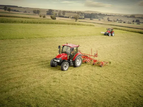 Rød traktor som arbeider på et grønt jorde med landbruksutstyr i kupert landskap.