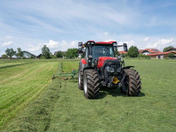 Rød traktor fra Vestrum-serien som raker gress på en grønn eng under blå himmel.