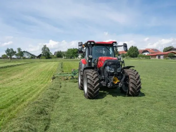 Rød traktor fra Vestrum-serien som raker gress på en grønn eng under blå himmel.