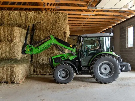 Grønn Deutz-Fahr 5D Keyline traktor som stabler store høyballer inne i en låve.
