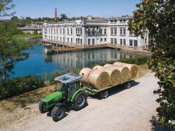 Grønn Deutz-Fahr 5D Keyline traktor transporterer store høyballer langs en elv med industribygg bak.
