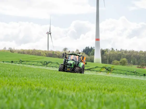 Grønn Deutz-Fahr Series 6.4 traktor som sprøyter en åker med vindmøller i bakgrunnen.