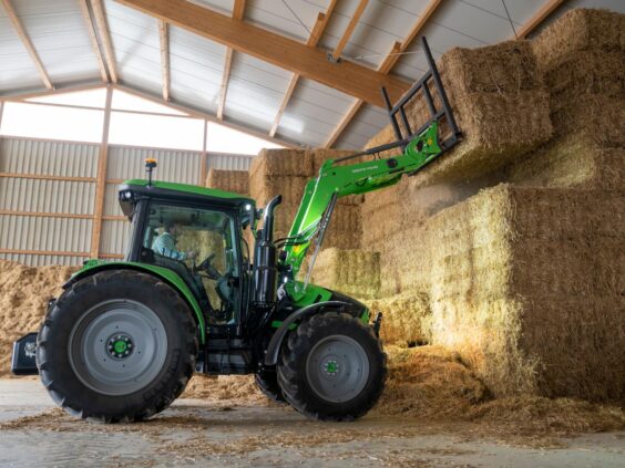 En grønn Deutz-Fahr Series 5 traktor som løfter store halmballer inne i en låve.