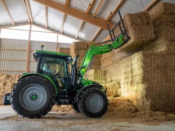 En grønn Deutz-Fahr Series 5 traktor som løfter store halmballer inne i en låve.