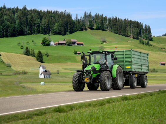 Grønn Deutz-Fahr Series 5 traktor med tilhenger på en landevei i vakkert kulturlandskap.