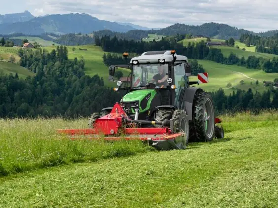 En Deutz-Fahr Series 5D traktor med rød frontslåmaskin som klipper gress i vakkert fjellandskap.