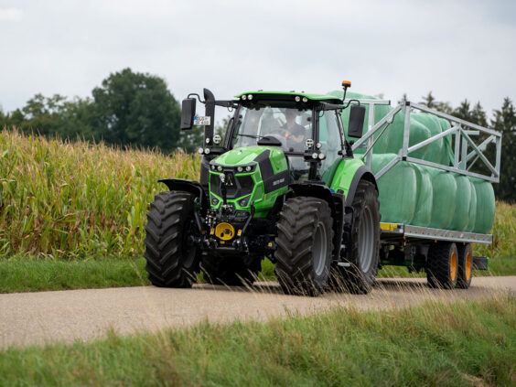 Grønn Deutz-Fahr Series 6 traktor som trekker en henger med rundballer langs en kornåker.