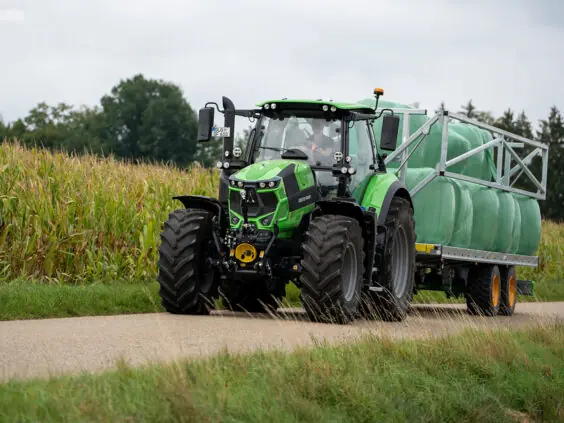 Grønn Deutz-Fahr Series 6 traktor som trekker en henger med rundballer langs en kornåker.