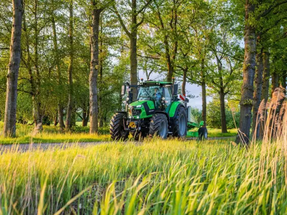 En grønn Deutz-Fahr Series 6.4 traktor kjører på en vei gjennom en frodig skog.