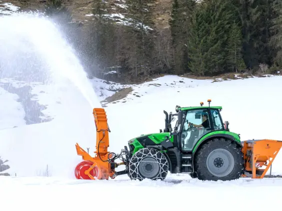 En grønn Deutz-Fahr Series 6.4 traktor med snøfreser i arbeid i et snødekt fjellandskap.
