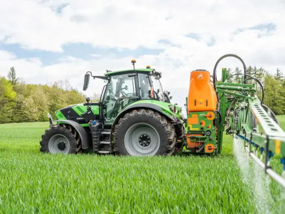 Grønn Deutz-Fahr Series 6.4 traktor som sprøyter en frodig åker under en skyete himmel.