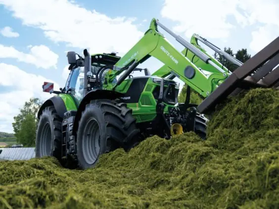 Grønn Deutz-Fahr Series 7 traktor med frontlaster som flytter gress på en gård.